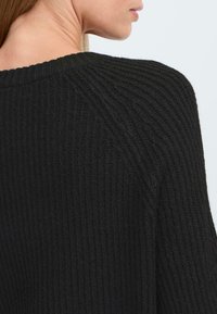 Pull en tricot noir avec une texture côtelée, doté d'un large décolleté et de coutures d'épaules abaissées, mettant en avant la douceur et la chaleur.
