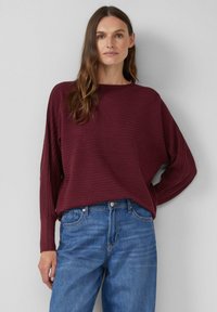 Bordowa bluzka z długim rękawem o prążkowanej fakturze i szerokim dekoltem w kształcie łódki, zestawiona z jasnoniebieskimi dżinsami. Minimalistyczny design z luźnym krojem.