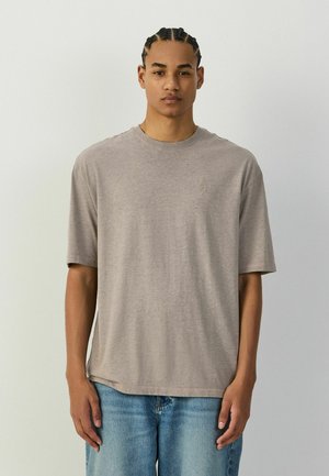 Junger Mann mit geflochtenem Haar, der ein einfaches beiges Kurzarm-T-Shirt und blaue Jeans trägt und vor einem hellgrauen Hintergrund steht.