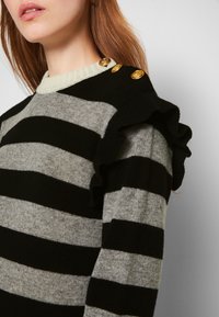 Femme portant un pull rayé noir et gris avec des détails à volants et trois boutons dorés sur l'épaule.