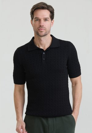 Rövid barna hajú férfi, fekete, rövid ujjú, kötött mintás pólóingben és zöld nadrágban, egy kézzel a zsebében áll.