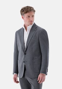 Grauer Wollanzug mit Revers, zwei Fronttaschen, Ein-Knopf-Verschluss und slim fit. Darunter trägt er ein weißes Hemd.