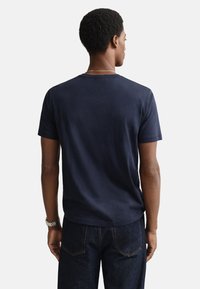 Mörkblå bomulls-T-shirt med rund hals och korta ärmar, visad bakifrån. Par mörka denimjeans med synlig söm.