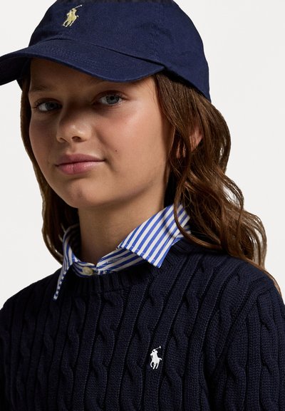 Pulôver de malha em cordão na cor azul-marinho, com logotipo branco bordado, sobre uma camisa com colarinho azul e branca com riscas, completado com um boné combinado.