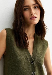 Gilet sans manches vert olive avec un décolleté en V, une texture douce et trois boutons noirs sur le devant.