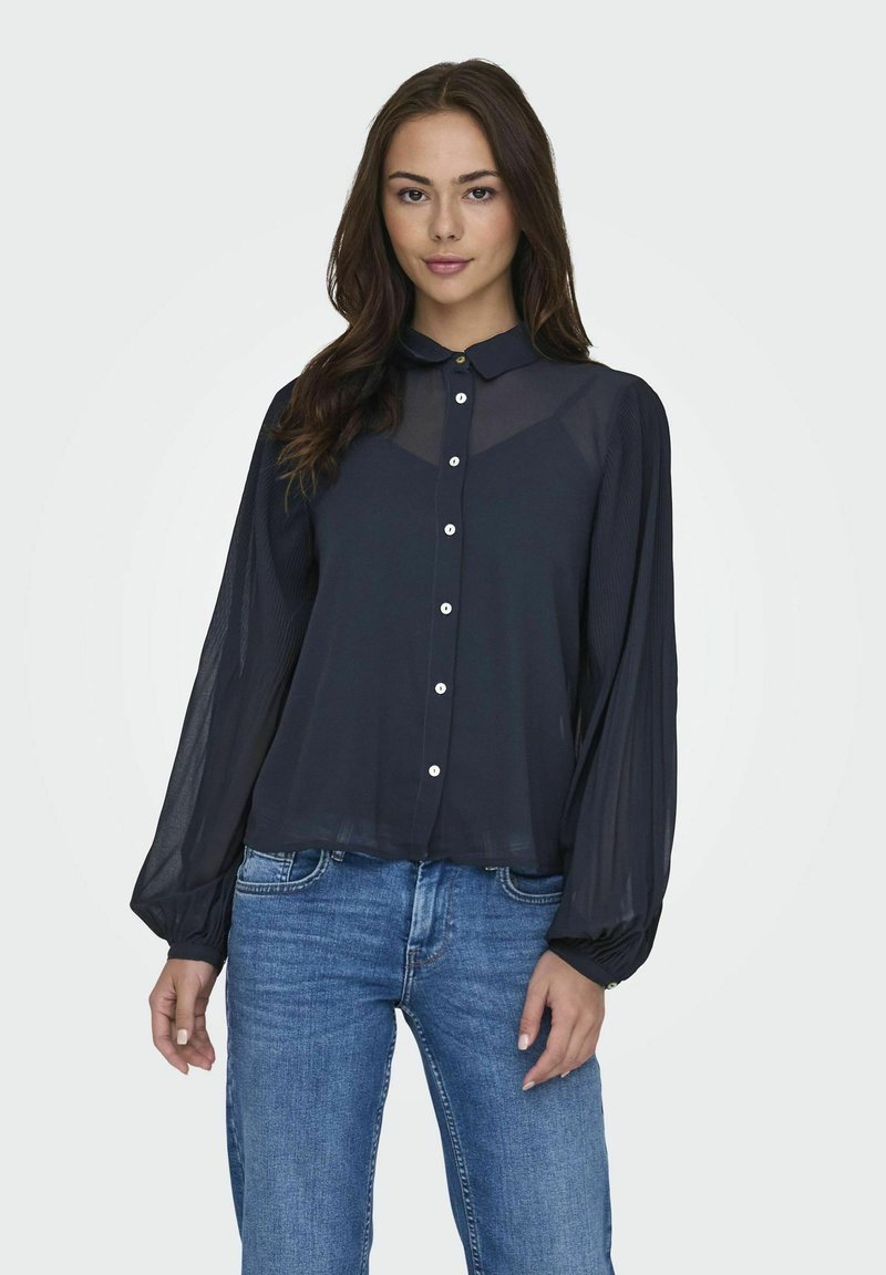 Blusa trasparente blu navy con collo a camicia, chiusura a bottoni e maniche lunghe plissettate, abbinata a jeans blu.