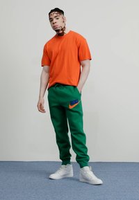 Orange bomullströja och gröna sweatpants med broderad blå och orange Nike-logotyp. Modellen har vita sneakers på en neutral bakgrund.