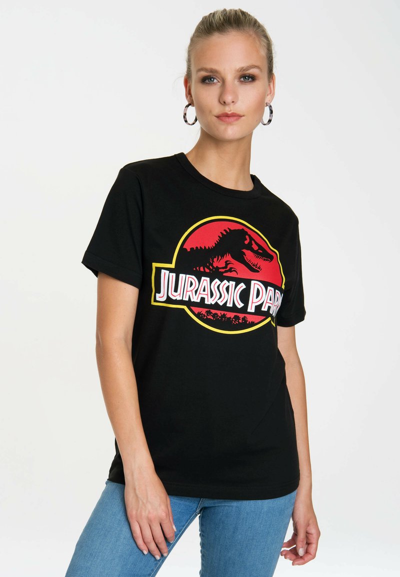 Schwarzes Baumwoll-T-Shirt mit einem roten und gelben Jurassic Park-Logo-Graphic. Kurzärmel mit Rundhalsausschnitt, kombiniert mit blauen Jeans.