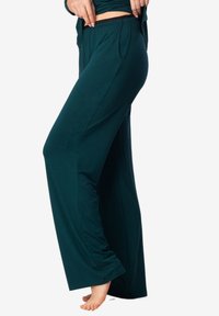 Turkusowe spodnie typu wide-leg wykonane z miękkiego materiału, z płaskim pasem, bocznymi kieszeniami i luźnym krojem. Model bez butów pokazany z profilu.