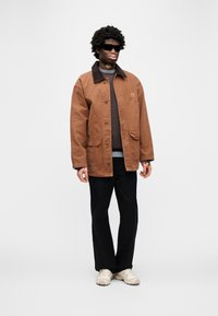 Καφέ καμβά μπουφάν με σκούρο γιακά, δύο μπροστινές τσέπες· φορεμένο πάνω από καφέ πουλόβερ και μαύρο παντελόνι, συνδυασμένο με ανοιχτόχρωμα παπούτσια.