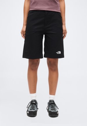 Person iført sorte The North Face-shorts, grå og sorte vandresko, hvide sokker og en lilla skjorte, der står på en ensfarvet baggrund.