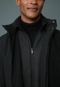 Chaqueta negra exterior con cuello alto, que cuenta con una cremallera y botones a presión. La capa interior tiene una textura con un diseño acolchado, ambas en negro.