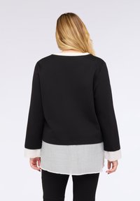 Černý top s dlouhými rukávy a kulatým výstřihem, vrstevnatý přes bílý, průhledný lem. Hladká textura a uvolněný střih zdůrazněné kontrastními manžetami.