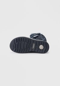 Sorte sneakerstøvel med en teksturert gummisåle, tre borrelåsremmer og en sirkulær logodetalj nær hælen. Myk fôr synlig på toppen.