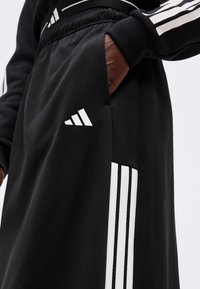 Χέρι στην τσέπη μαύρου φόρμας Adidas με λευκή λεπτομέρεια τριών ρίγες και λογότυπο Adidas στο μηρό.