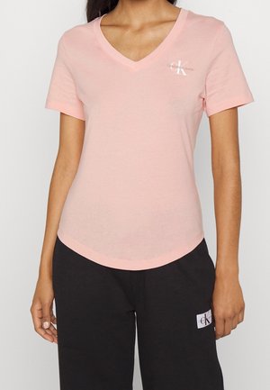 Frau trägt ein hellrosa V-Ausschnitt-T-Shirt mit kurzen Ärmeln und kleinem Calvin Klein Logo sowie schwarze Hose mit Calvin Klein Aufnäher.