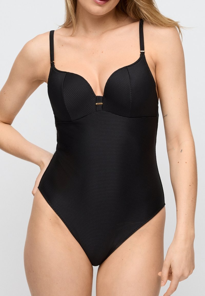 Vrouw draagt een nauwsluitend zwart geribbeld eendelig badpak met verstelbare schouderbandjes, staand met één hand op haar heup tegen een effen achtergrond.
