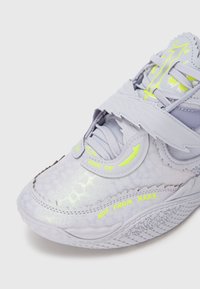 Světle šedá atletická obuv s texturovaným, reflexním svrškem, zdobená neonově zelenými detaily a zapínáním na pásky. Výrazný zakřivený design podrážky.