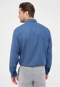 Camicia a maniche lunghe da uomo in blu scuro, tessuto testurizzato, con colletto, polsini con bottone e dettagli sullo yoke posteriore, abbinata a pantaloni grigio chiaro.