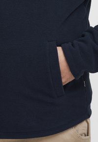 Pullover in pile blu navy caratterizzato da una texture morbida, tasche laterali e orlo dritto. Il tessuto sembra caldo e leggero.