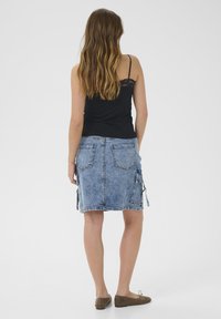 Denimrock mit einer verblassten blauen Waschung, mittellang, ausgestattet mit zwei seitlichen Reißverschlusstaschen und zwei aufgesetzten Gesäßtaschen. Kombiniert mit einem schwarzen Tanktop.