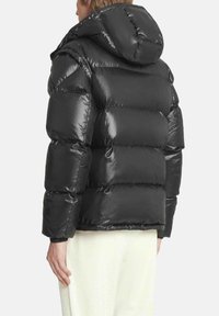 Giacca imbottita nera con finitura lucida, dotata di cappuccio e design boxy trapuntato. Le maniche sono leggermente affusolate; il materiale è liscio e isolante.