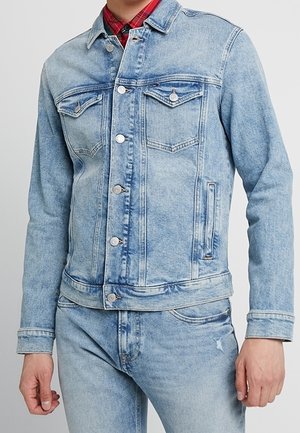 Hombre con chaqueta vaquera azul claro y jeans con botones visibles, bolsillos en el pecho y una camisa de cuadros roja debajo.