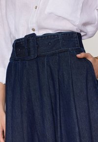 Denimrok met hoge taille, brede riem met een vierkante gesp, geplooid ontwerp en diepblauwe kleur, gecombineerd met een wit overhemd met kraag.