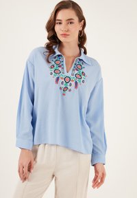 Hellbluse mit langen Ärmeln und Schlitzkragen, verziert mit buntem Blumenstickerei und einem strukturierten Stoff.