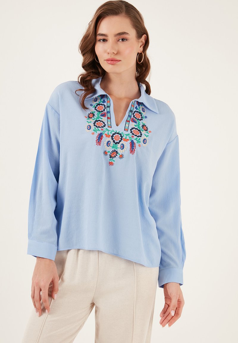 Hellbluse mit langen Ärmeln und Schlitzkragen, verziert mit buntem Blumenstickerei und einem strukturierten Stoff.