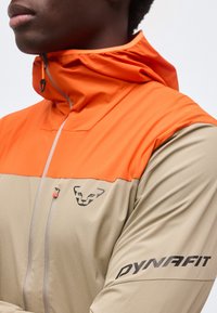 Κοντινό πλάνο ενός ατόμου που φοράει μπεζ και πορτοκαλί μπουφάν Dynafit με κουκούλα, τσεπάκι με φερμουάρ στο στήθος και λογότυπο Dynafit στο μανίκι.