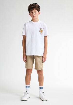 T-shirt en coton blanc avec impression "Key West", shorts beiges et baskets blanches avec des accents verts. Des chaussettes rayées bleues complètent la tenue.