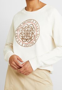 Sudadera crema con un gráfico circular marrón que presenta diseños intrincados y texto. Las manos del modelo muestran un agarre relajado, con puños acanalados.