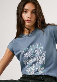 Zila kokvilnas t-krekls ar kapučiem, ar ziedu rakstu un baltu tekstu "PEPE JEANS LONDON", izvietots uz gaiši zaļas fona.