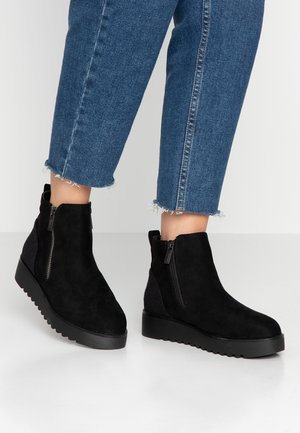 Bottines en suède noir avec des zips latéraux, présentant un panneau gris texturé sur le talon et une semelle noire épaisse et striée.
