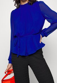 Blue, long-sleeve blouse with a high neck, pleated design, and a tie waist. Paired with textured black pants and a red handbag.