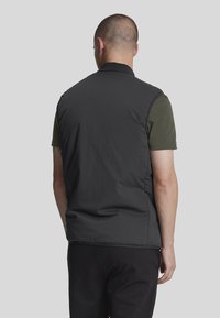 Gilet nero senza maniche con una texture liscia e un colletto alto, indossato sopra una camicia verde, caratterizzato da un design minimalista e senza hardware visibile.