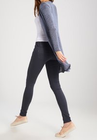 Lehký pletený kardigan v jemné modré barvě, otevřený střih s třásňovým lemem, v kombinaci s tmavými úzkými džíny a béžovými casual botami.