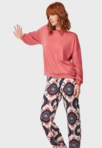 Sudadera rosa de corte relajado, combinada con pantalones de crema con patrones que presentan diseños de ojos en negro, rojo y amarillo. Tela de textura suave.