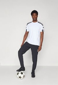 Ung man i vit Adidas-tröja och svarta byxor står med ena foten på en svart och vit stjärnmönstrad fotboll framför en neutral bakgrund.