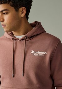 Ruda hoodie su raišteliais, ant priekio esantis baltas logotipas "Manhattan Cask & Craft". Lygus audinio tekstūra ir standartinė apranga.