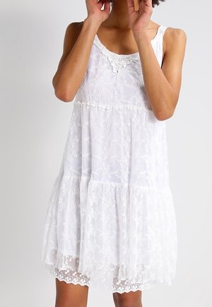 Femme portant une robe blanche sans manches en dentelle avec des motifs floraux et une jupe à étages, visible des épaules jusqu'au milieu des cuisses sur un fond uni.