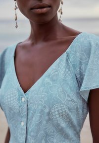 Vestido de tela azul claro con estampado de flores y piñas, con escote en V, mangas cortas y detalle de botones en la parte delantera.