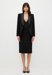 Musta mukautettu puku, jossa on istuva takki, jossa on satiinikaulukset, syvä V-pääntie ja polven mittainen kynähame. Slim fit -malli, jossa on vähäiset yksityiskohdat.