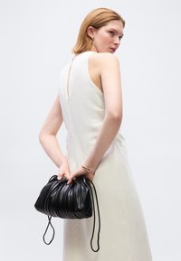 Γυναίκα με άσπρο φόρεμα χωρίς μανίκια κρατά μια μαύρη τσάντα με πιέτες πίσω από την πλάτη της, σε λευκό φόντο.