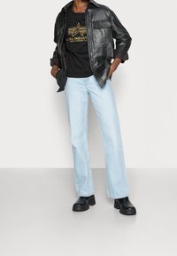 Veste en cuir noire portée sur un sweat-shirt noir avec un logo doré, associée à un jean large bleu clair et des bottines noires épaisses.