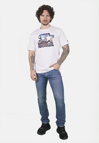 T-shirt bianca con un design grafico colorato che presenta una stella e del testo. Abbinata a jeans in denim blu e sneakers nere, su uno sfondo chiaro.