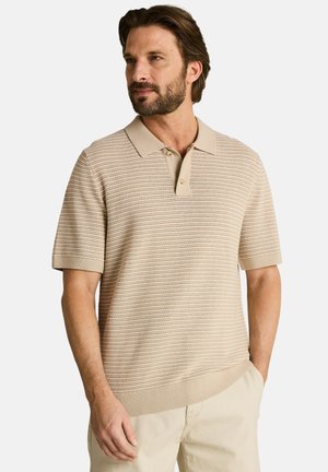 Homme portant un polo en maille texturée beige à manches courtes et un pantalon clair, debout avec une main dans la poche, regardant vers la gauche.