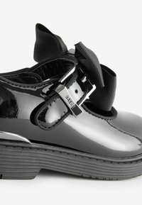 Schwarzer Lacklederschuh mit runder Zehenform, strukturiertem Gummisohle und einer großen schwarzen Schleifenakzent, gesichert durch eine Metallniete.