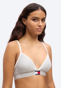 Νεαρή γυναίκα με μακριά σκούρα μαλλιά, φορώντας ένα ανοιχτό γκρι bralette Tommy Hilfiger με λεπτές τιράντες, μπροστά σε απλό φόντο.